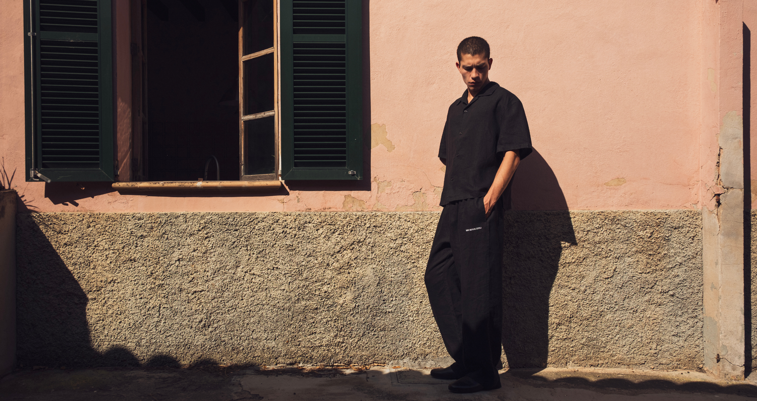 A man wearing a black short-sleeve button-up shirt and matching black trousers stands against a pink stucco building wall with dark green shutters, casting a long shadow.