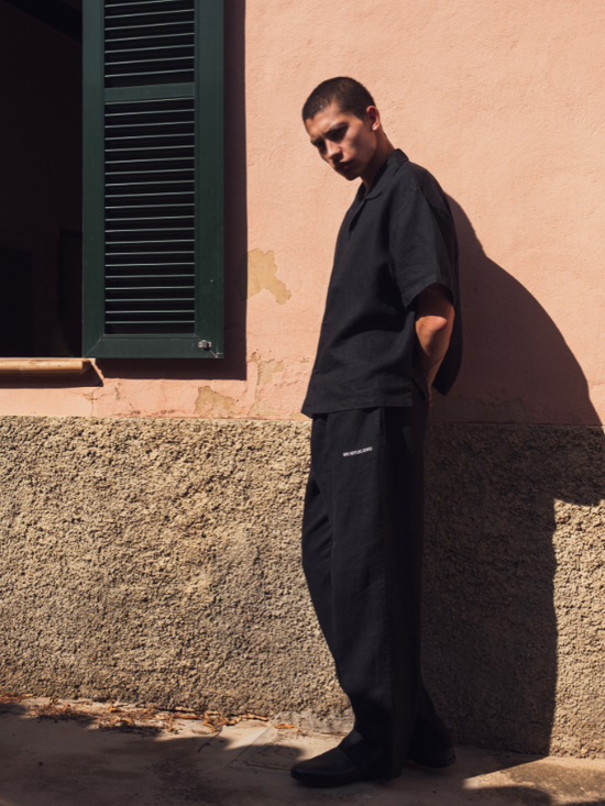 A man wearing a black short-sleeve collared shirt and matching black trousers with a small white logo on the thigh, leaning against a textured stucco wall next to a dark green shutter.